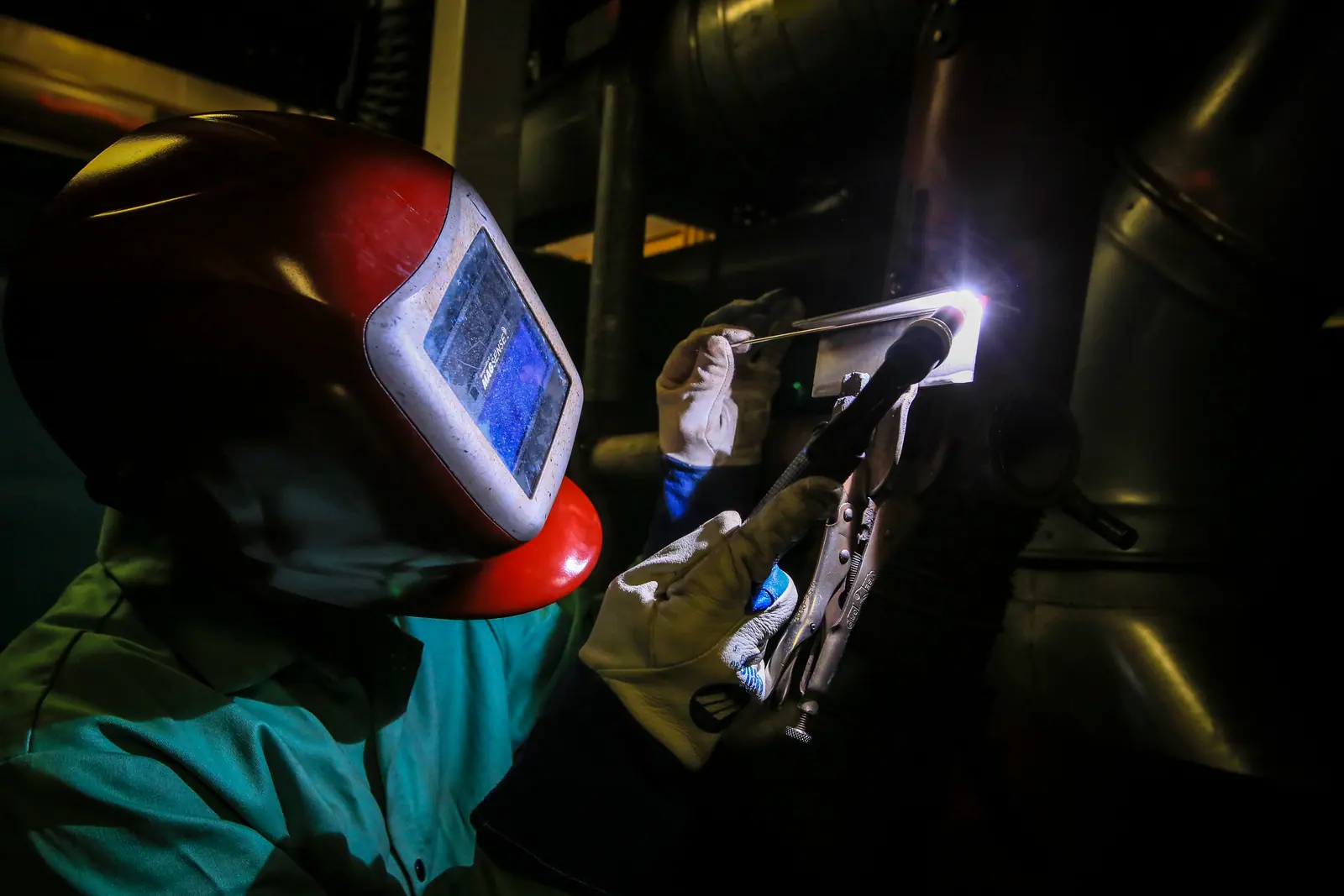 A welder in a red helmet and leather gloves running a TIG torch on a metal plate, the arc glowing white